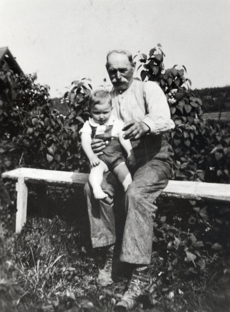 Emanuel Johansson på Helgö gård sitter på en bänk med barnbarnet Björn i knäet.  Björn är son till Seth Helgöstam och hans hustru Hanna