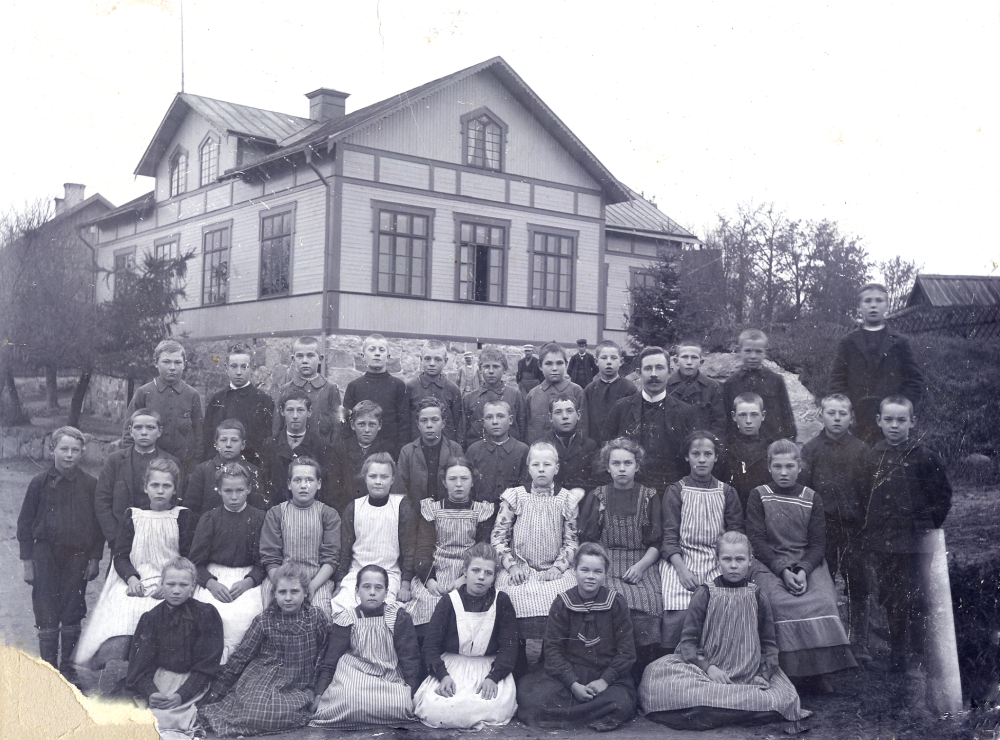 Kyrkskolan i Össeby-Garn med läraren Bernhard Wiberg, förmodligen 1908.  I nedre raden nr 3 fr v med randigt förkläde sitter Anna Elisabet Kjellberg, gift Forsberg, som bodde på Toftesta gård i Össeby-Garn