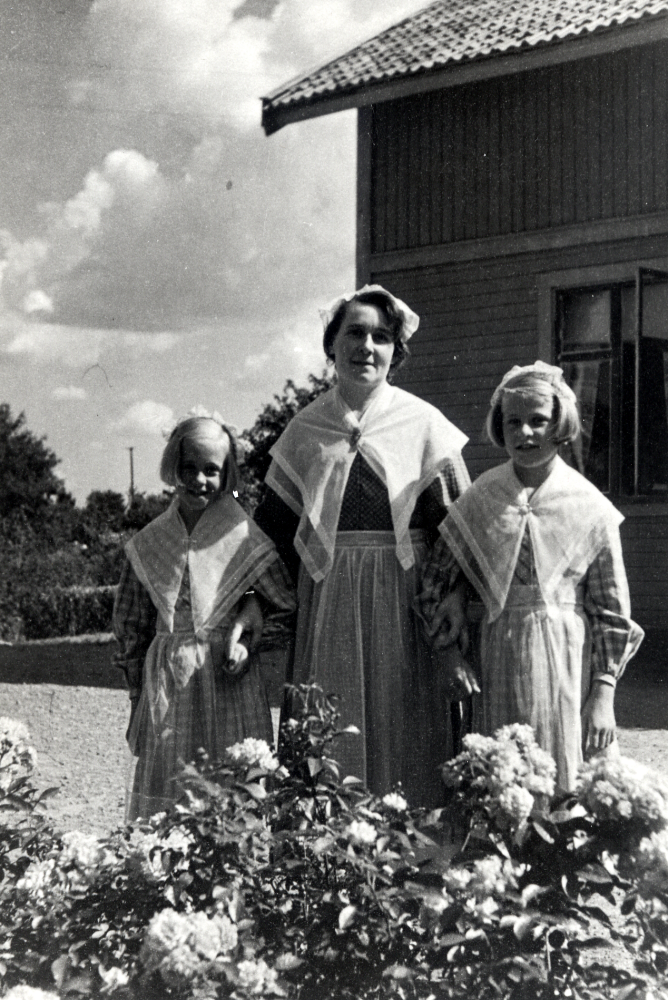 Märta Karlsson, hustru till Sven Karlsson, arrendator på Väsby gård, med döttrarna Ulla t v och Marianne, alla klädda i upplandsdräkten.  Foto vid rundeln där flaggstången står, i bakgrunden gårdens huvudbyggnad.  Familjen är på väg till Bondens dag på Skansen i Stockholm.  Fotografen är brorsbarn till gammelfastrarna Jenny och Hanna på Väsby gård