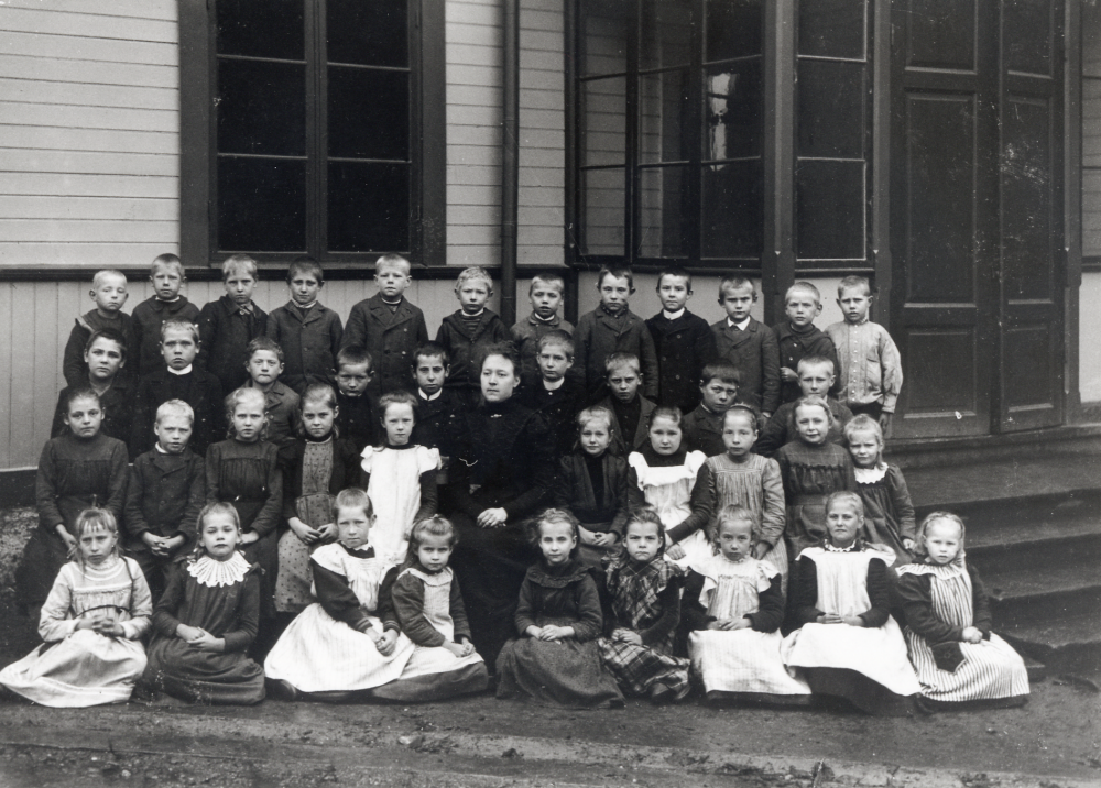 Skolbarnen tillsammans med lärarinnan utanför kyrkskolan i Lilla Garn.  Flickan i vitt förkläde t h om lärarinnan heter Selina Andersson, född 1896 och moster till Sven Dahlbeck