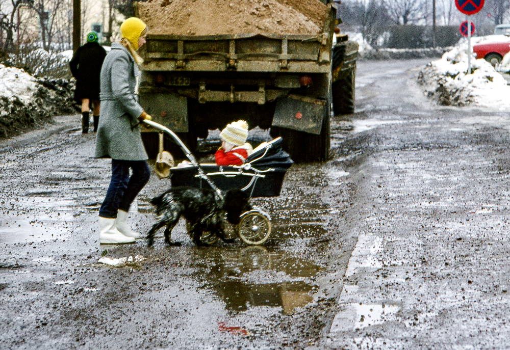 Förälder med barn i barnvagn och en hund passerar Banvägen. I bakgrunden en lastbil fullastad med sand.
Bildserie: Vallentuna på 1960-talet.