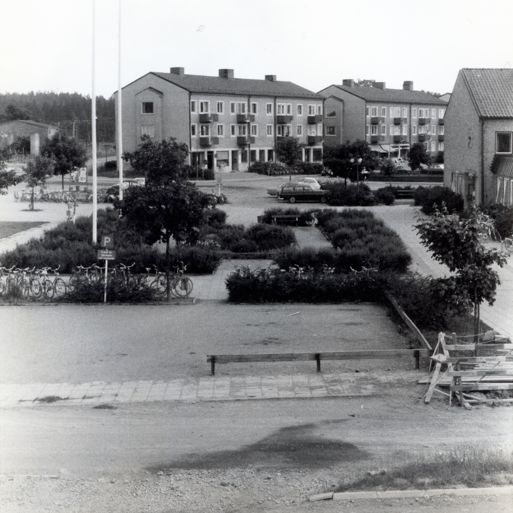 Vallentuna centrum tidigt 1970-tal, före augusti 1972. Vy över parkeringen och parken framför det gamla kommunalhuset sett från Allévägen där ett mindre vägarbete pågår till höger i bild. Längst bort ligger bostadshusen på Centralvägen med butiker i markplan. 
