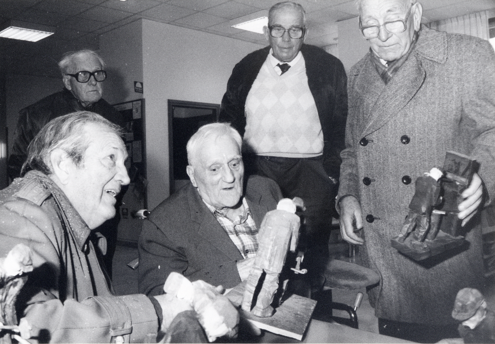 Utställningen Smia i Vallentunasalen får besök av den gamle smeden K S Karlsson, Sme-Kalle. Runt den gamle smeden fr v Halvar Näslund, okänd, Sven Jansson  "Plåt-Janne " och Gustav Nilsson.  Utställningen Smia presenterade bysmedjorna i Uppland, sammanställd av Hans Lustig i samarbete med Uplandsmuseet.  Hans Lustig har också gjort de olika träfigurerna i utställningen