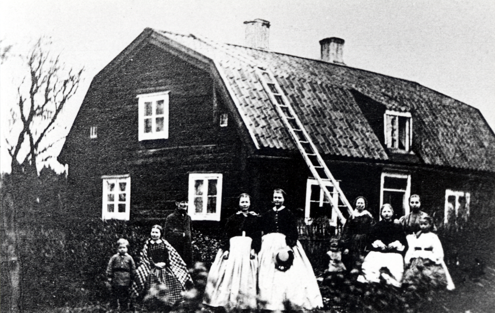 Björkby komministerboställe i Vallentuna.  Personer fr v Martin Svansson 1857-1909, Augusta Hjertqvist 1822-1882 gift med Arvid Svansson,,  Arvid Svansson 1809-1893, blev komminister i Vallentuna 1849 och bosatt på Björkby till 1870 då han blev kyrkoherde i Skånela, hans sista ord var: "Jag går från mycket elände till stor härlighet!". Hanna Svansson 1854-1926, mor till Julia som omtalas nedan,  Augustina Svansson 1850-1942, Elisabeth Svansson 1861-1945, en guvernant, samt tre okända personer.  Huset byggdes 1750.  Omkring 1860 var Björkby på 4 mantal, lönen för komministern 350 riksdaler.  Av pastoratet fick han 6 och av församlingen 24 riksdaler, vidare fick han i utsäde 30 lass hö, men inget bete och ingen skog ingick.  1999 är huset starkt förfallet.  På detta fotografi är båda takhalvorna brutna, 1999 är norra takhalvan sadeltak och den södra bruten.  Framför huset står en ek som kallas Julias ek. Förmodligen planterad som dopgåva till Julia Lundberg, dotterdotter till Arvid Svansson