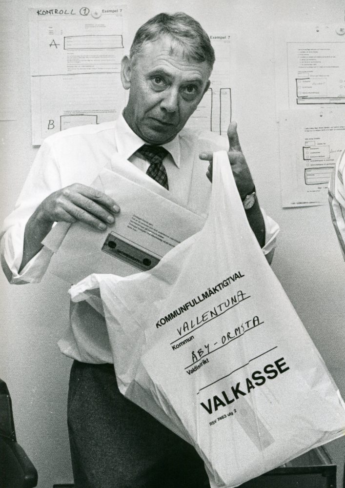 Val 1988. En man stoppar ner kuvert i en plastkasse märkt "Kommunfullmäktigeval, Kommun Vallentuna, Valdistrikt Åby-Ormsta, Valkasse. Namnuppgift saknas.