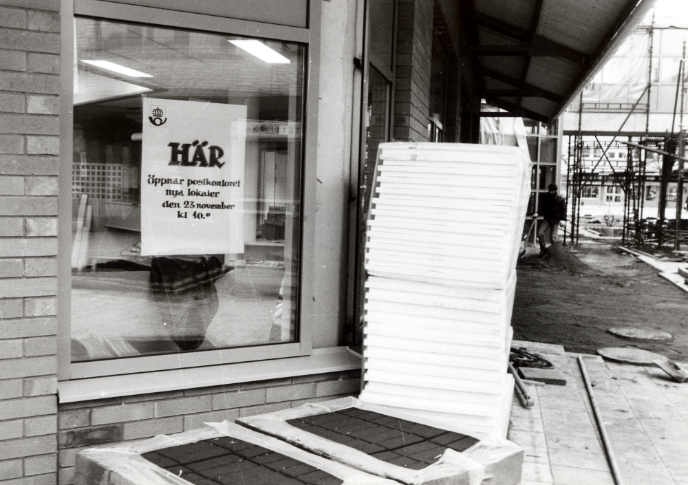 Vallentuna kommun bygger nytt kommunalhus.  I bottenvåningen flyttar bl a posten in i större lokaler från Tunahuset.  På skylten i fönstret står ' Här öppnar postkontoret nya lokaler den 23 november kl 10.00'