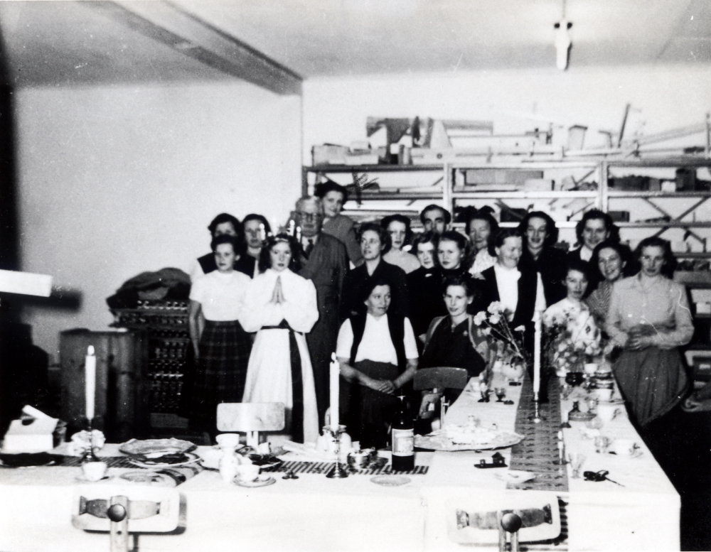 Personal vid L M Ericsson i Vallentuna firar Lucia.  L M Ericsson hade lokaler i Svedjevägens industriområde.  Sittande i mitten klädd i väst är Tyra Karlsson.  Längst bak bakom mannen i glasögon står Vivi Danred, senare gift med Hilding Eriksson, tågmästare på Roslagsbanan