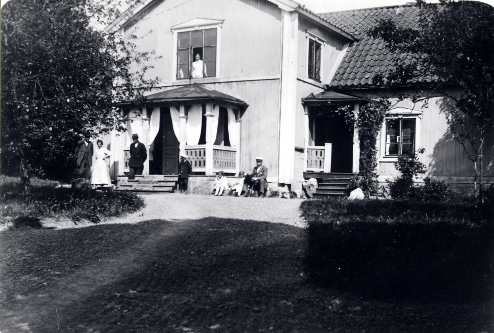 Framför Husby gård står fr v Gottfrid Andersson, hans hustru Hanna Andersson, Helge Andersson, Gottfrids och Hannas barn Nils Andersson, Inga Andersson och Elsa Andersson samt gårdens ägare Theodor Gustafson . I fönstret står Julia Berglund