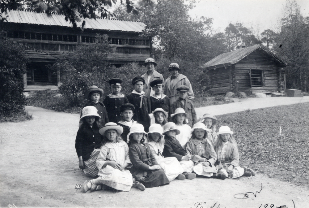 Lärarinnorna  Nanny Skoglund  t v och  Rut Nilsson  tillsammans med skolbarnen från  Össeby-Garns kyrkskola i första raden.
Tillägg 2020-10-05: Svar om platsen från Skansens personal som tror att bilden är tagen vid Kaplansloftet och Hackstugan vid Orsakullen på Skansen.