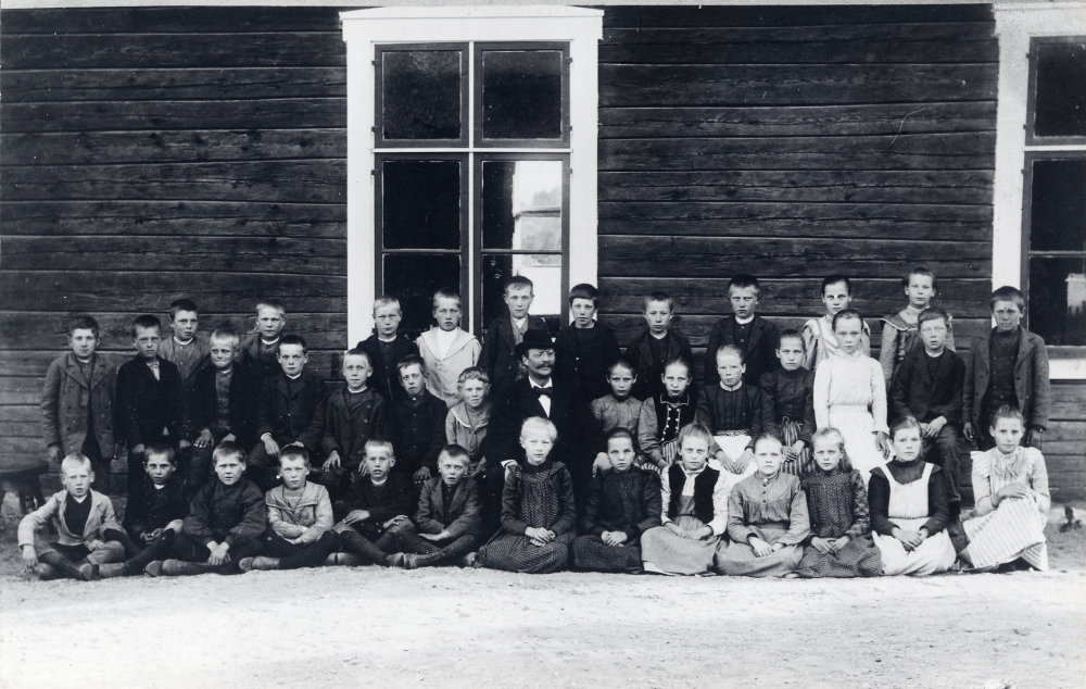 Läraren Hjalmar Zetterlund tillsammans med skolbarnen utanför Markim-Orkesta skola. Något t v om läraren i bakre raden står David Lust (sedemera antagit släktnamnet Jansson). Flickan stående längst t h är Maria Karlsson gift Gehlin. Zetterlund var initiativtagare till bildandet av en skytteförening. I nästan varje socken startades en sådan vid sekelskiftet. K H Zetterlund var lärare i socknen 1885-1906. Förutom studentexamen och lärarexamen hade han utbildning i trädgårdsskötsel och trädplantering, bokbinderi och vaccination