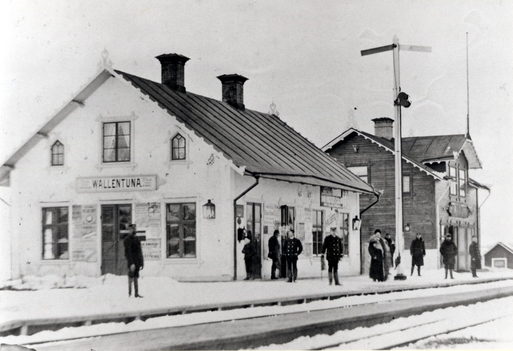 Vallentuna järnvägsstation med järnvägsrestauranten Flugan t h.  Vid stationshuset står en semafor.  På plattformen står järnvägspersonal i uniform och bl a en man med pälsfodrad långrock