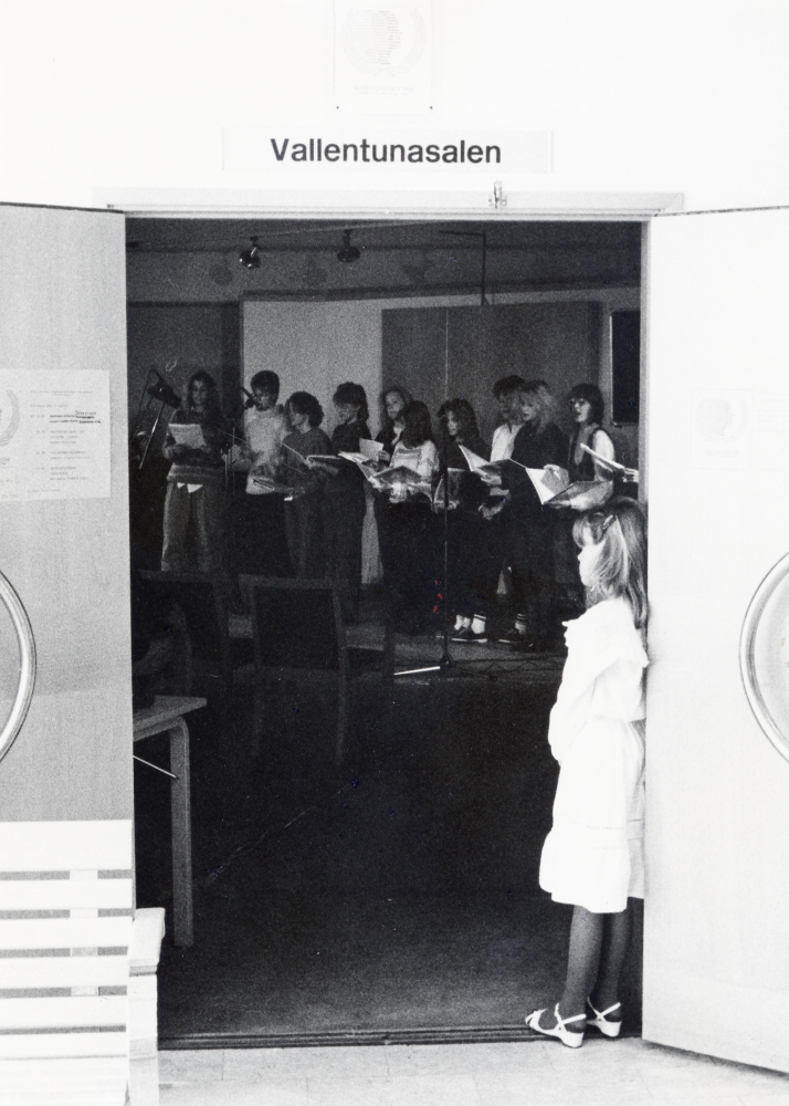 I Vallentunasalen pågår ofta program och utställningar arrangerade av Vallentuna kulturnämnd.  Salen ligger i anslutning till Vallentuna bibliotek och foajé.  Här står en liten flicka i dörröppningen när en ungdomskör sjunger från podiet inne i salen. Tema: 1985 - Världsungdomsåret. Plats på scen: Ungdomsföreningar och -grupper