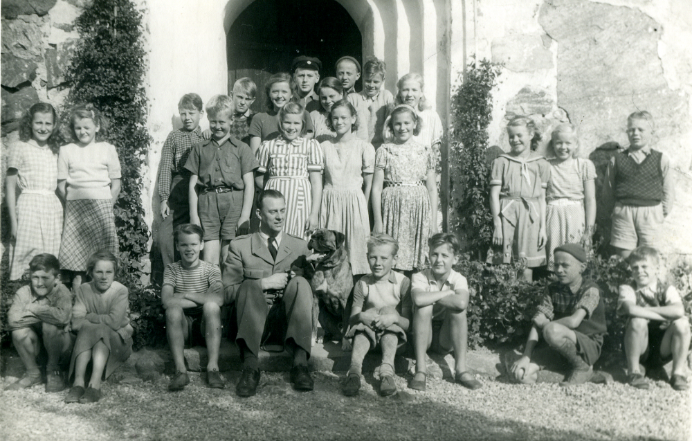 Läraren Gösta Sahlin med skolbarnen i klasserna 5-7 i Össby-Garns kyrkskola utanför Össby-Garns kyrka.  Längst bak i porten står fr v Olle Pettersson (Almén) Brottby, Ingvor Eriksson Ekeby, Lasse Rödin Ösby, Barbro Pettersson Brottby, Arne Johansson Gillberga, Yngve Alstermark Kristineberg och Ragna Alstermark Ösby.  Mittenraden fr v Marianne Lundberg, Gunilla Wettervik Brottby, Hasse Stjernholm Hagalund, Gösta Johansson Hakunge, Astrid Gustavsson Skeppartorp, Margareta Balk Lilla Garn, Eva Britt Persson Skrattby, Britt Nordström Lövhamra, Ulla Jansson Ösby och Lasse Gustavsson Sjötorp. Sittande fr v Björn Larsson Karby?, Iris Larsson Karby?, Stig Jansson Ösby, läraren och hunden Tello, Gösta Andersson Pälsen, Max Hultgren Sundby, Rune Johansson Gillberga och Bengt Öberg Karby