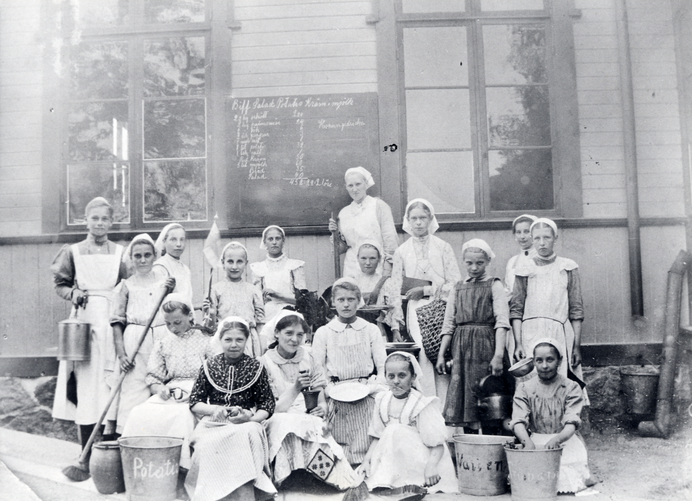 Skolkök i Össeby-Garns skola Lärarinnan i bildens mitt är okänd, t v om henne Frida Bergqvist med mjölkflaskan Elna Karlén mörk blus har Helga Bergqvist t v om henne Ester Rydin Kortet var länge okänd, först efter publicering i DN Nordost 810305, gavs dessa uppgifter. I bakgrunden en tavla med uppgifter om den måltid som lagades till ett pris av 21 öre/portion