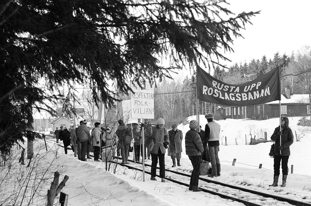 Protester inför hot om nedläggning av Roslagsbanan; Föreningen Rädda Roslagsbanan protesterar inför hotet av nedläggning av järnvägen. Trafiken Kårsta-Rimbo upphörde i januari 1981.
I bakgrunden skymtar Kårsta stationshus och t.h. det gamla affärshuset Österlid, sedan 1969 ombyggt till bostad för familjen Staff.