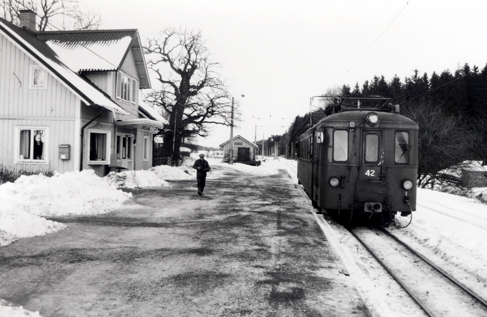 Kårsta järnvägsstation.  Vid perrongen står motorvagn 42 som pendlar mellan Kårsta och Lindholmen.  På plattformen sandar banarbetaren Bernt Eriksson, som arbetar som spårsynare och banvakt