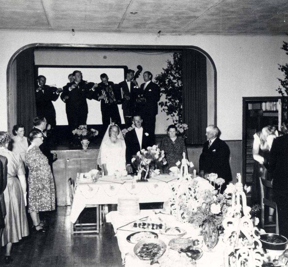 Ingegärd Eliasson och Stig Öhmans bröllopsfest i Ekskogens bygdegård i Kårsta.  Runt brudparet står fr v Berit Gustavsson, Stina Jansson, Gärda Eliasson och  prästen Paul Sjöberg T h står fr v Alma Öhman, brudgummens mor och Einar Eliasson brudens far.  Musiker på scenen är fr v Enar Lundqvist,  Erik Holmberg, Åke Mattsson, Harry Franzon, okänd och Sven Wahlström.  På bordet i förgrunden står smörgårdsbordet dukat och två  bröllopskrokaner pryder bordet