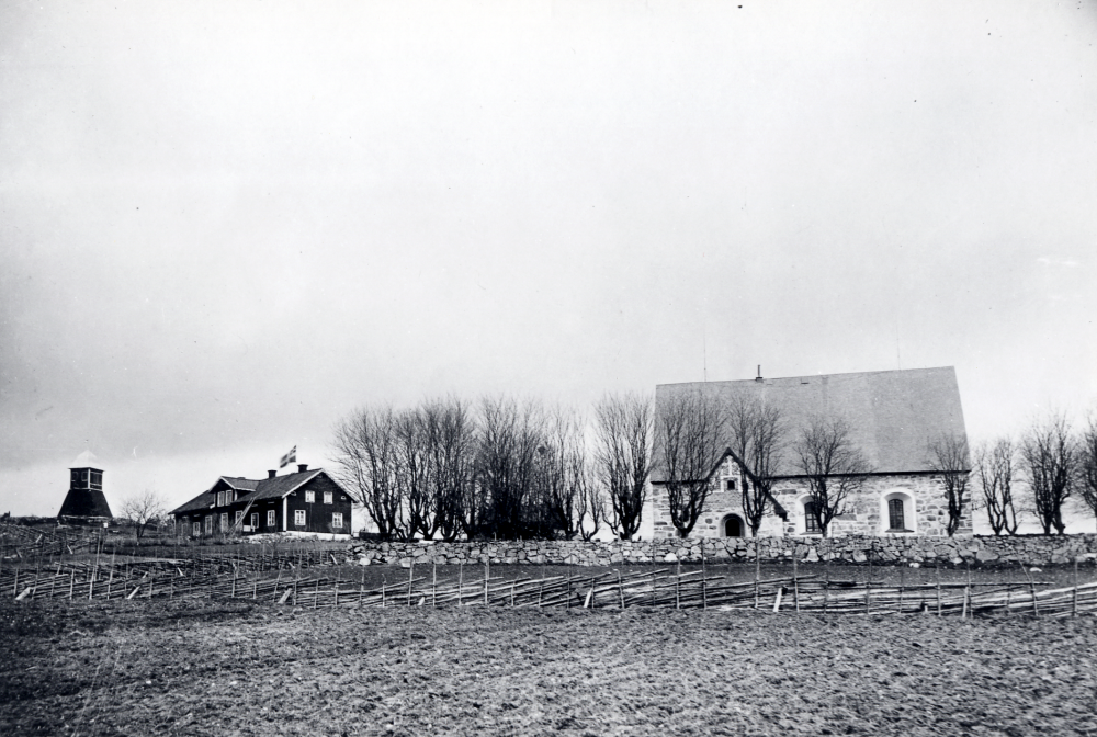 Kårsta kyrka och Kårsta folkskola sedda från söder. T v ligger klockstapeln. I förgrunden plöjd åkermark och en gärdsgård