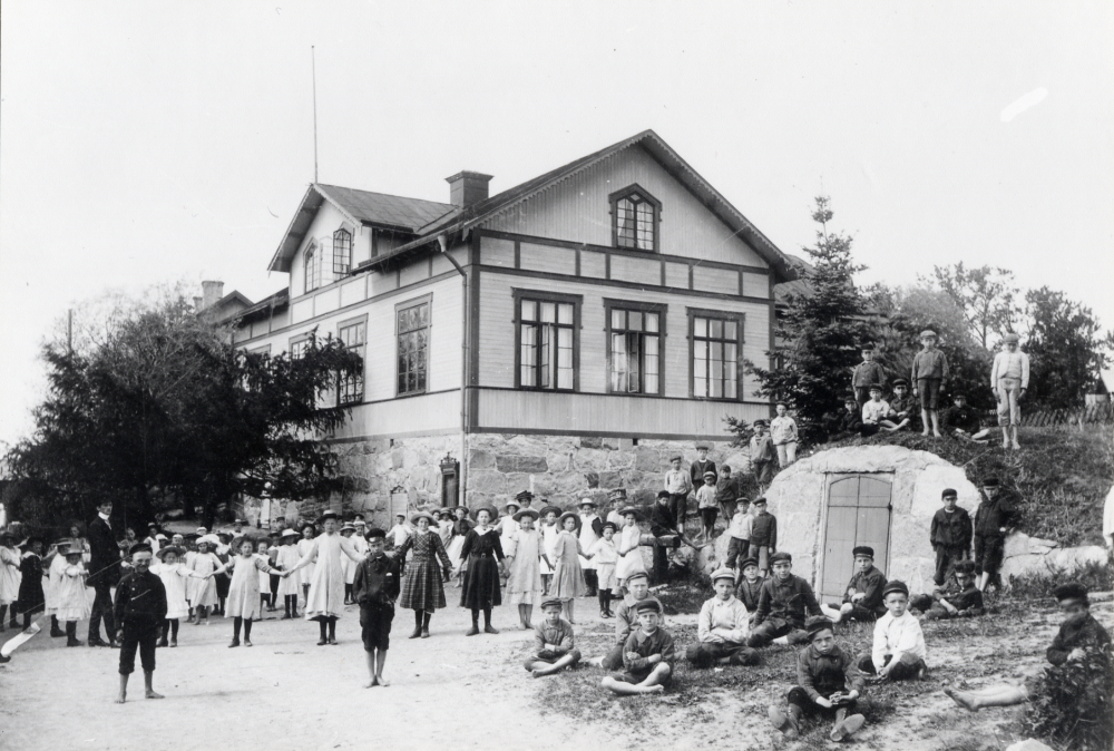 Lärarna  Bernhard Wiberg?   Nanny Skoglund  och  Mathilda Linde  tillsammans med skolbarnen utanför  Össeby-Garns kyrkskola  Lärarinnorna står vid skolan gavel iklädda stora hattar. T h syns dörrarna till en  jordkällare  Barnen är vackert grupperade på vägen utanför skolan