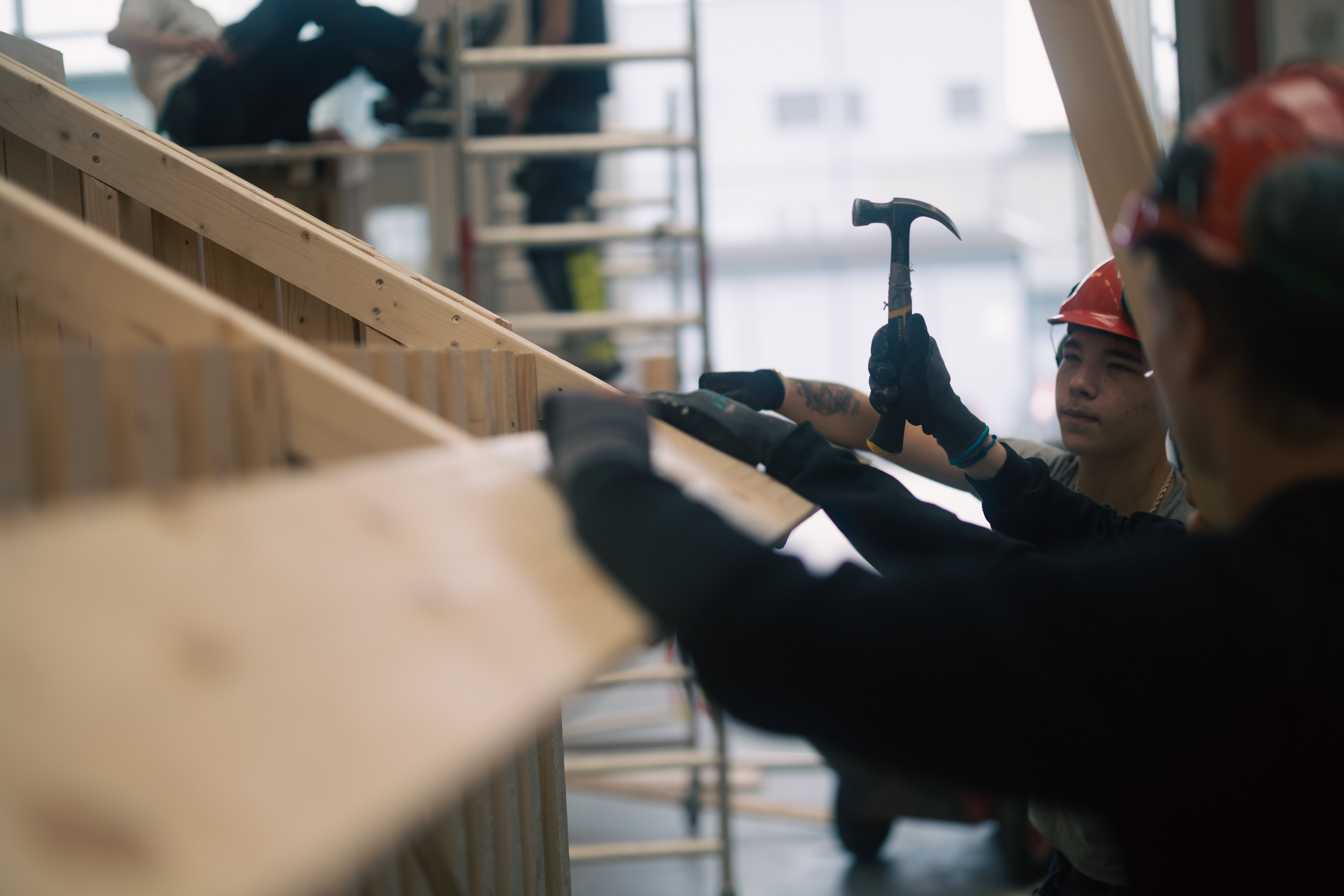 Två elever från bygg- och anläggningsprogrammet placerar och hamrar fast en bräda som ska agera tak till ett modulhus.