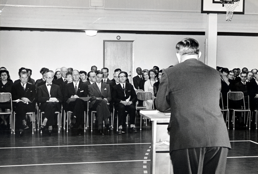 Skolstyrelsens  och  kommunalfullmäktiges  ordförande  Bertil Tillborg  talar vid  invigningen  av  Vallentuna sporthall  vid  Hjälmsta skola.  I främre raden sitter fr v rektor  Josef Andersson,  riksidrottsförbundents representanter  Eskil Tidén och  Nordenskiöld,  länsskoleinspektör  Helldén  och ordförande i länsskolnämnden  Sven Öhrn.   Andra raden från mittgången mot dörren från höger sitter kontraktsprost Sven Vängby  och folkskollärare  Bernhard Bengtsson