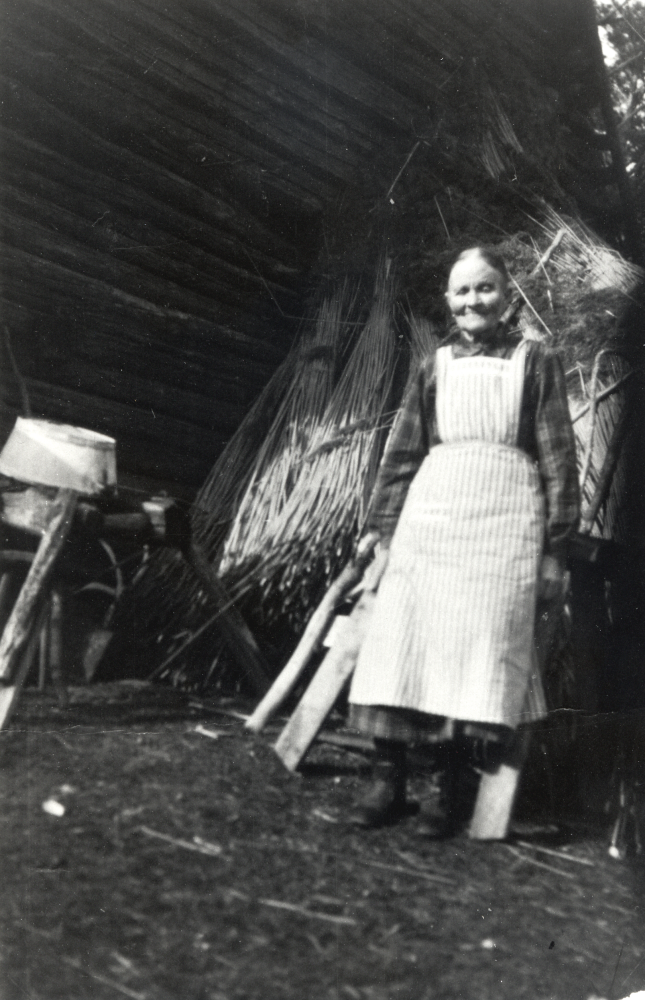 Selma Andersdotter i Fågelsången vid uthuset med vassknippen på tork mot husväggen.  Selma ägde ett stort markområde vid Fågelsången