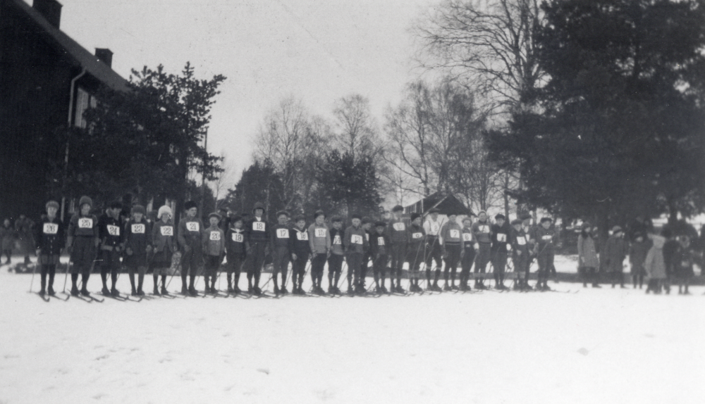 Den årliga skidtävlingen vid Hjälmsta skola var en uppskattad händelse varje vinter.  Skolbarnen med nummerlappar på bröstet har ställt upp på skolgården med Röda skolan i bakgrunden t v.  De barn som inga skidor hade, fick  genom skolans försorg
