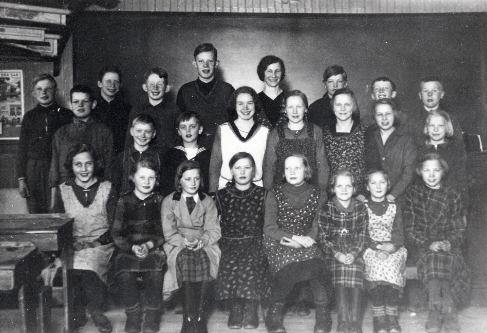 Lärarinnan Elna Andersson tillsammans med skolbarnen i klassrummet i Angarns storskola. Bakre raden fr v Rune Berggren, Gösta Hjelm, Henry Eriksson, Tord Johansson-Tordmar, Yngve Jansson, Bertil Jansson och Viktor ?.  Mellanraden fr v Evert Andersson, Nils Sjöman, Knut   Sjöman, Svea Sjöman, Astrid Grandin, Margareta Andersson, Britt ? och Majsy Jansson.  Nedersta raden fr v Rakel Axén, Gelly Andersson, Ulla Johansson, Astrid Lindersson, Gunnel Johansson, Valborg Nilsson, Aina Lindersson och Ester? Svensson