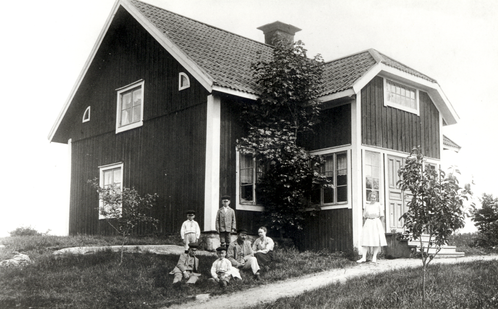 Pelsen i Ösby, Össeby-Garn. Vid husgaveln sitter John, (Johan Alfred) född 1883 och hustrun Hulda Dahlbeck född 1883 med sönerna sittande framför föräldrarna fr v Georg född 1909 och Valter född 1915, stående bakom fr v Gunnar född 1916 och Sigurd född 1913.  Vid trappan står pigan Tyra Alstermark född 1906 senare gift Sundström