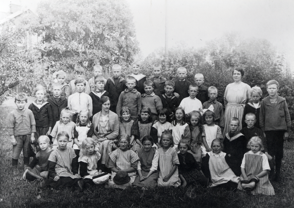 Utanför Angarns skola sitter lärare och skolbarn. Övre raden fr v Olof Eriksson Åsta, okänd, Stig Andersson Lillåsta, okänd, Artur, Bertil Lindecrantz Rocksta, okänd, och Sixten Jansson Lövlund. Andra raden stående fr v Sven Åkersten Ö Båtmanstorp, Evert Lindström Alby, Sten Nilsson Rörby, Helmer Andersson Lycktan Haga, Sixten Lindström Alby, Nils Jansson Lövlund, Set Nilsson Rörby,  Nils Hellberg, Veda, Arne Dahl Rävsta, Harry Högberg Rocksta, lärarinnan Ida Solem, okänd och Anton Lindecrantz Rocksta.  Rad 3 sittande på en bänk Gunvor Johansson Rävsta, Cissi Olsson Åsta, lärarinnan Elna Andersson, Karin Andersson Lycktan Haga, Hildegard (Knopanie) Brissman Örsta, Valborg Lindström Alby, Elsa Olsson Åsta, Herta Nilsson Rörby och Inez Eriksson Åsta. Rad 4 fr v okänd, Lilly Lindecrantz, Gullan Dahl Dragontorpet Kinnekulle, Lisa, Tyra Logran Damsta, Ingrid Johansson Rävsta, Hildur Nilsson Rörby, Karin Gustavsson Kinnekulle, Einar Andersson Veda, Oskar Jansson Lövlund och Gunhild Törnkvist Damsta