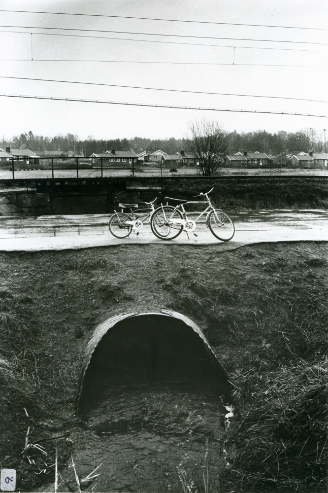 Vårflod i bäcken vid Banvägen. Ett par cyklar med "limpor" till sadel står parkerade ovanför bäcken som går genom Vallentuna. "Farlig lekplats" har fotografen skrivit bredvid fotot. 
•Fotografiet ingår i bildserien ”Vardagsliv i Vallentuna kommun” av Rachael Gough-Azmier & Gunilla Blomé, Haga Studios HB.