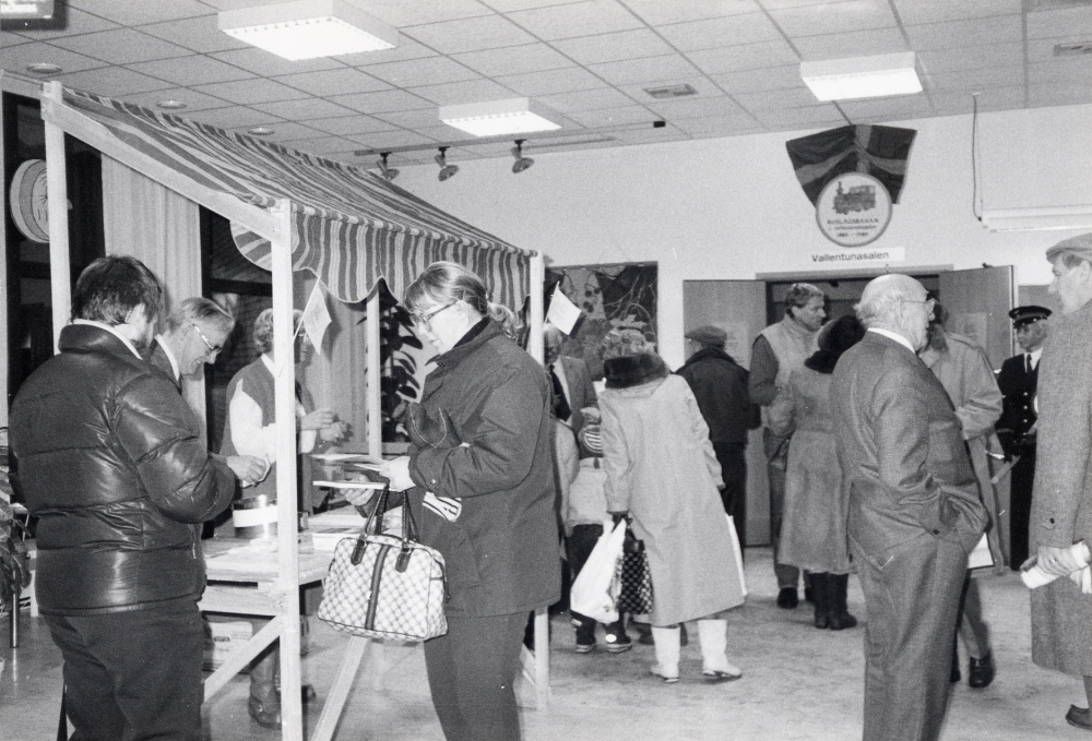 Roslagsbanan fyller 100 år på riktigt den 19 december 1985 och firas med bl a utställning i Vallentunasalen och glögg m m i foajén utanför Vallentuna bibliotek. Hembygdsföreningarna längs Roslagasbanan och SRJ firade 100-års jubileet den 21 september 1985 med ångloksdraget tåg m m. Vid ståndet i foajén serverar Sven Skoog t v och Gunhild Nylén Hillbo besökarna glögg. Med rutig bag står Susanna Stubbendorff och tittar i boken Roslagsbanan och bygden utgiven i samband med jubileet. I bildens mitt med skidväst står Anders Fröderberg, t h samtalar Gustav Nilsson med Gunnar Welander. Vid den ljusa flaggani bildens mitt skymtar Gustaf Åkerblom.