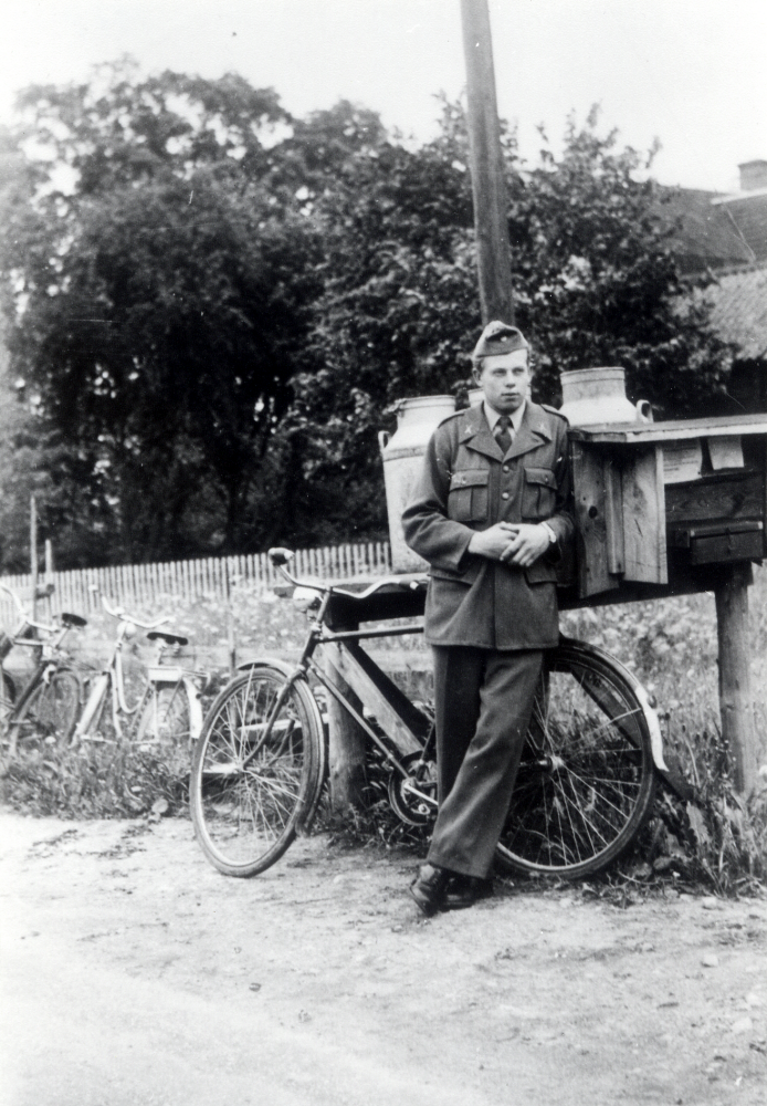 Sixten Helgöstam från Vreda i Frösunda, soldat vid krigsslutet 1946, står med sin cykel lutad mot ett mjölkbord med två mjölkflaskor.  I bakgrunden Frösunda prästgård?