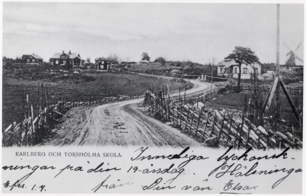 Vägen förbi Torsholma skola. Torsholma skola ligger t h om vägen. Det största röd-vita huset t v om vägen är Karlbergs handelsbod. Rättelse 2021-01-28: Enligt nya uppgifter är det Eriksborgs kvarn som syns längst till höger, inte Torslunds kvarn som tidigare angavs. Samma bild som 133. Vykort på Karlberg och Torsholma skola med handskriven hälsning.