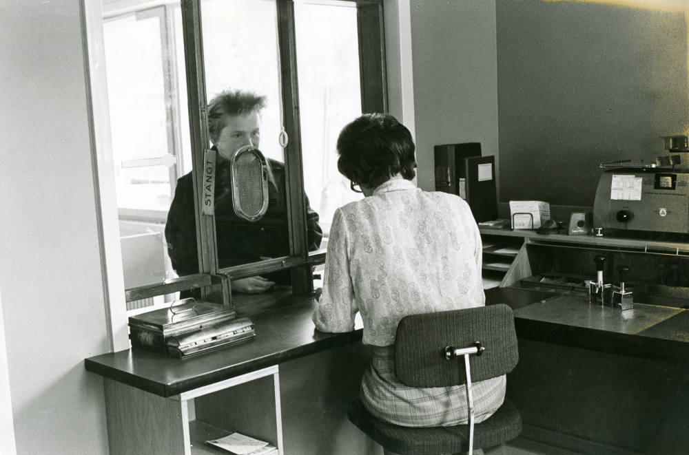 Det nya postkontoret i Kårsta har öppet en timma om dagen. Postens föreståndare fru Hewer sitter bakom disken och tar emot en kund.
•Fotografiet ingår i bildserien ”Vardagsliv i Vallentuna kommun” av Rachael Gough-Azmier & Gunilla Blomé, Haga Studios HB.
