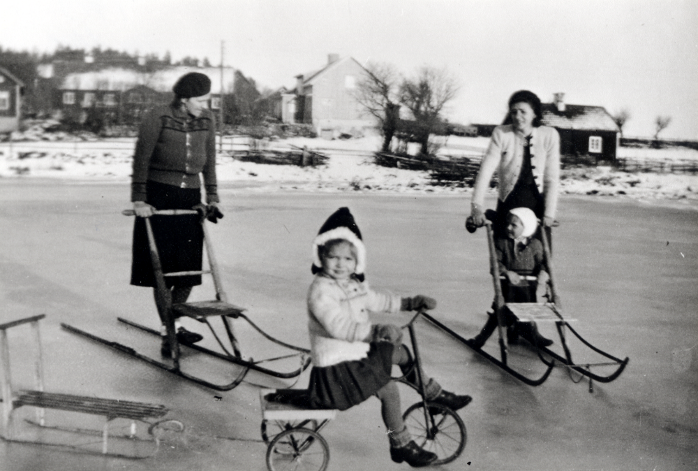 Vinter vid Helgö gård. På Helgösjöns is åker t v Alice Helgöstam sparkstötting, brorsdottern Inga Helgöstam trampar en trehjuling med en kälke fastknuten baktill och Margit Andersson (f Helgöstam) har också en sparkstötting med brorsonen Kjell på sitsen.  Barnen Inga och Kjell är barn till Bengt och Sonja Helgöstam på Helgö gård. I bakgrunden syns gårdens byggnader