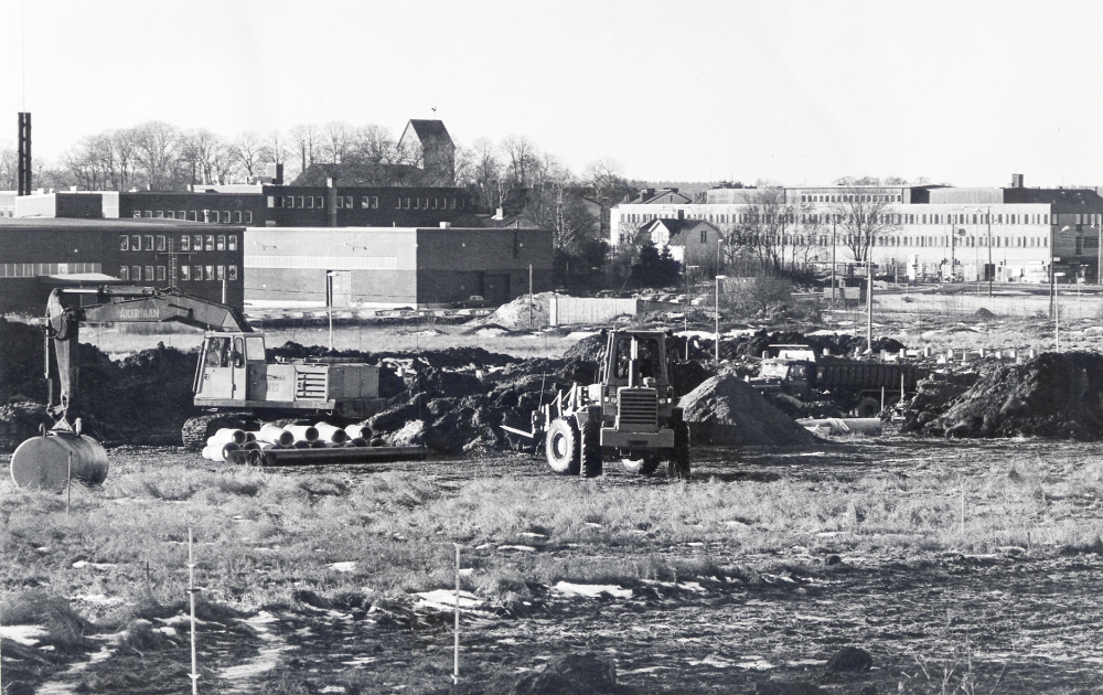 Schaktningsarbete  inför utbyggnaden av  Åby gärde  T v  Tellus Maksin  samt i bildens mitt  Tunahuset.  I bakgrunden syns  Vallentuna kyrka  I förgrunden en  grävskopa  och en  lastare. Lastaren står i ett blivande kvarter på Takpannevägen med adress Takpannevägen 111 – 193 (Taxizon 5).