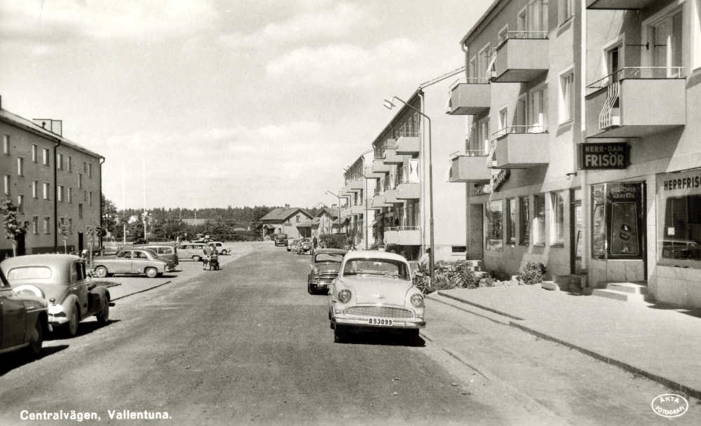 Centralvägen, Vallentunas första affärsgata, med flerfamiljshus och affärslokaler som byggdes i början av 1950-talet. Till höger finns en frisersalong, skylten visar att det är både herr- och damfrisör. Bredvid ligger Konsum snabbköp som senare blev lokal för Vallentuna varuhus. I bakgrunden mellan husen skymtar Vallentuna järnvägsstation med stationshuset till höger och till vänster järnvägsrestaurangen Flugan. En Opel Rekord står parkerad på Centralvägens högra sida (enligt uppgiftslämnaren kom bilmodellen ut 1956 i denna version). På andra sidan går Elsie Westlund med barnen Lars och Tommy och Kjell i barnvagnen.