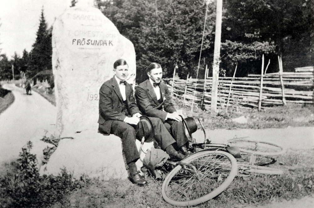 Vägvisare av sten vid Skrattbacken i Markim.  Framför stenen sitter t v Georg Åkerberg och t h Sven Åkerberg.  Framför de unga pojkarna ligger deras cyklar.  Texten på stenen lyder ovanifrån:  T. SKÅNELLA.  T. ORKESTA OCH FRÖSUNDA K .  Stenen restes 1920 av  Patron Teodor Gustafson från Husby gård i Markim