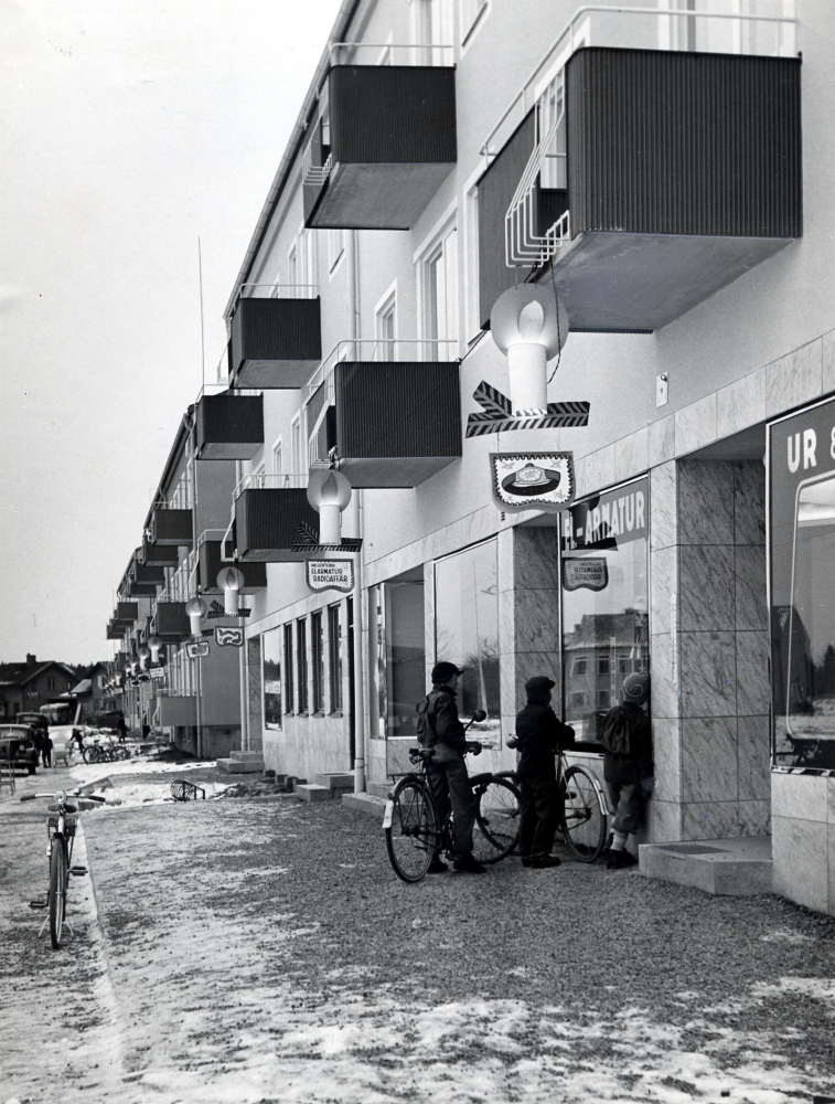Affärernas julskyltning på fasaden vid Centralvägen, Vallentunas första affärsgata, med flerfamiljshus och affärslokaler som byggdes i början av 1950-talet. Till höger ligger Vallentuna El-Armatur och Radioaffär samt Ur. Tre barn tittar in i skyltfönstret.  Fasaden på Kommunalhuset vid torget speglas i fönsterglaset. I bakgrunden till vänster skymtar  Vallentuna järnvägsstation  med  stationshuset och järnvägsrestaurangen Flugan.