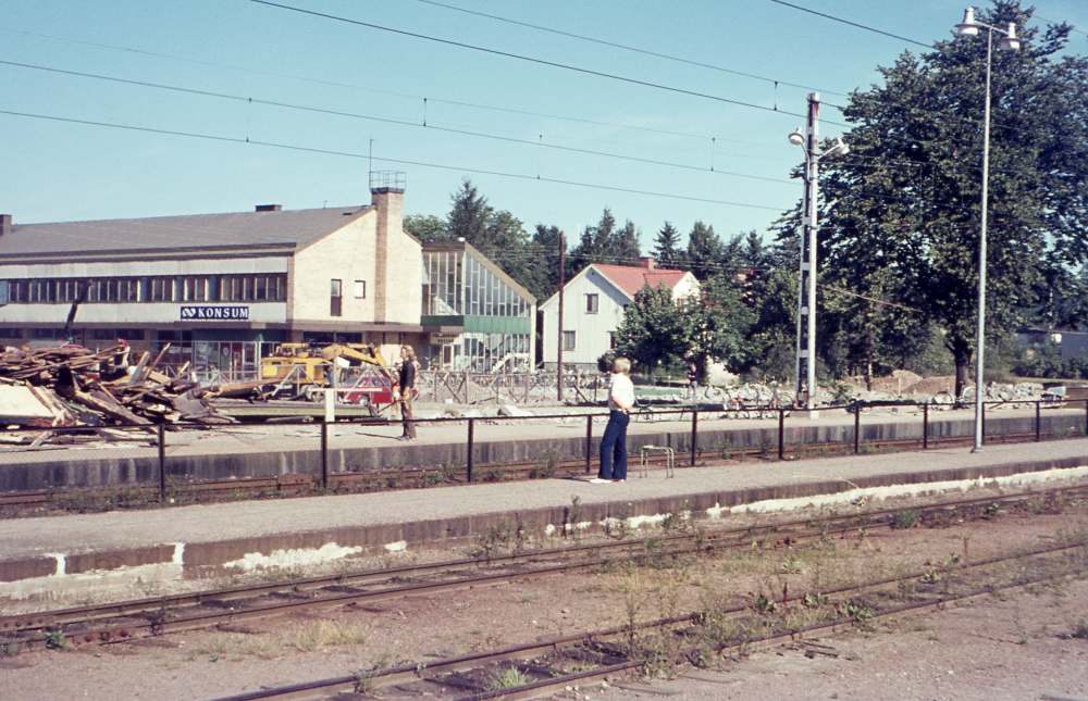 Rivning av Vallentuna stationshus pågår. En hög av bråte syns till vänster. I bakgrunden ligger Torghuset med Konsum, beläget vid korsningen Allévägen / Banvägen.