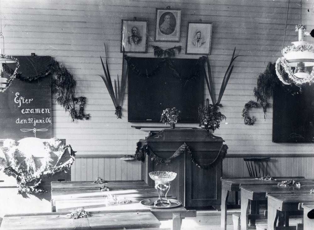 Frösunda skola  "Efter  Examen".  Skolsalen är pyntat efter skolavslutningen och på bordet står blommor och en glasskål, möjligen en present som läraren Zackeus Carlsson fått som examenspresent av skolbarnen