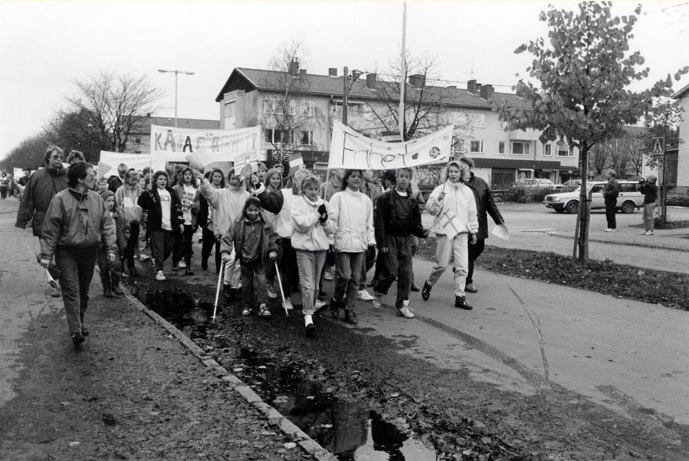 Barn från Ekebyskolan tågar till Kommunalhusparken på FN-dagen 24 oktober.  Här går de på Allévägen med sina banderoller. De har stora knippen med vita ballonger som de senare skall släppa upp i luften för fred i världen