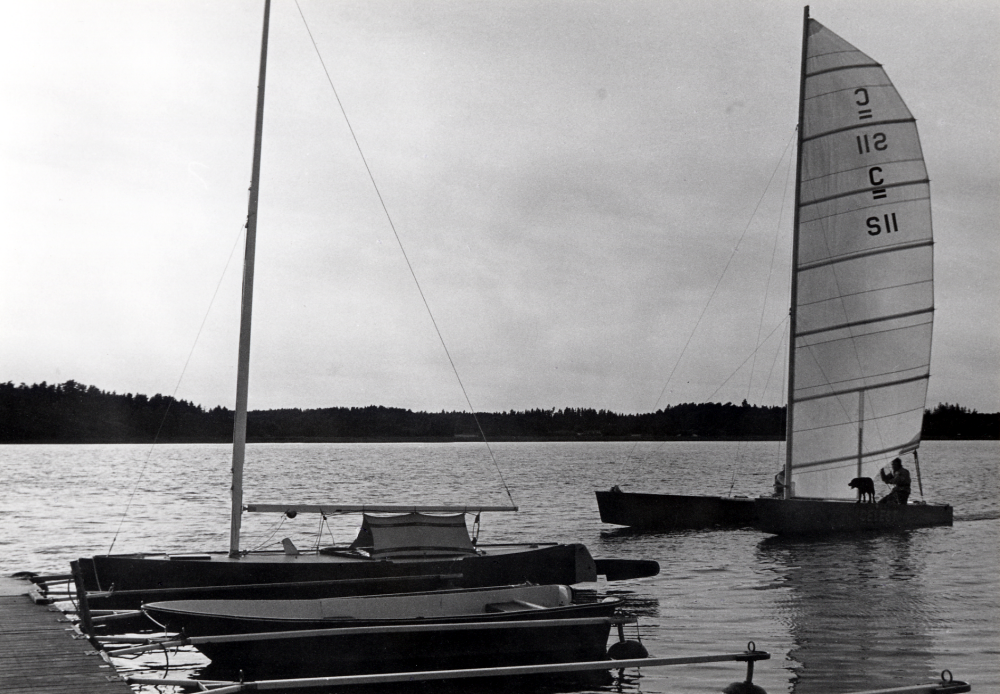 En segelbåt och en roddbåt vid kommunens båtbrygga vid Gustavs udde i Bällsta i Vallentunasjön. En segelcatamaran med två personer ombord samt en hund närmar sig bryggan