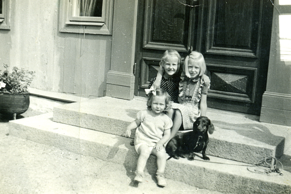 På trappan till Backa gårds manngårdsbyggnad sitter Elna Karlsson, Kerstin Lindgren och Anders Lindgren. Anders har på grund av sitt lockiga hår klätts i klänning av flickorna. Kerstin och Anders var barn till Backa gårds ägare Erik och Siri Lindgren.