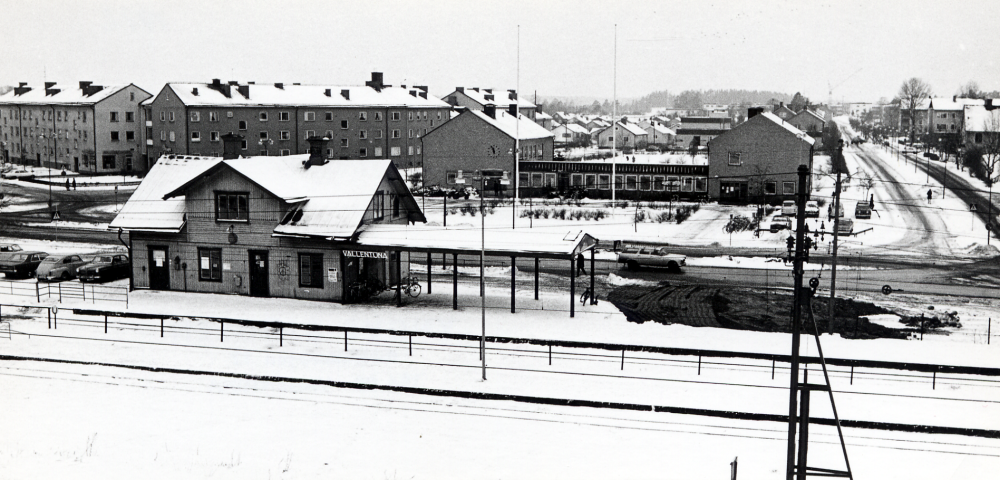 Vallentuna järnvägsstation med Vallentuna kommunalhus i bakgrunden. T h syns bostadshuses vid Skördevägen och Centralvägen T h syns Allévägen Fotograferat före byggstarten av Tunahuset framför kommunalhuset. Bredvid järnvägsstationen syns en grop där järnvägsrestauranten Flugan legat