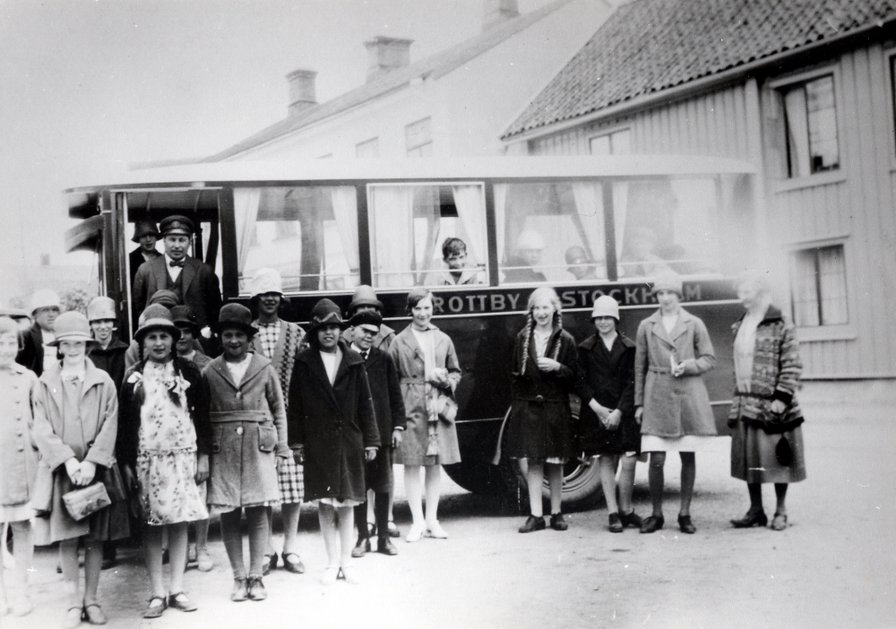 Buss märkt Brottby-Stockholm på skolresa, troligen till Mariefred. I bussdörren står chauffören Ture Pettersson, längst t h står lärarinnan Elna Andersson. Vissa barn är kända fr v: flickan med flätor Inga-Lisa Eriksson, i rutig klänning Lilly Lindecrantz, pojke i vegamössa Harry Johansson, pojke i bussen Karl-Olof Eriksson, ljus flicka i flätor Sissi Olsson, i ljus kappa Inez Eriksson