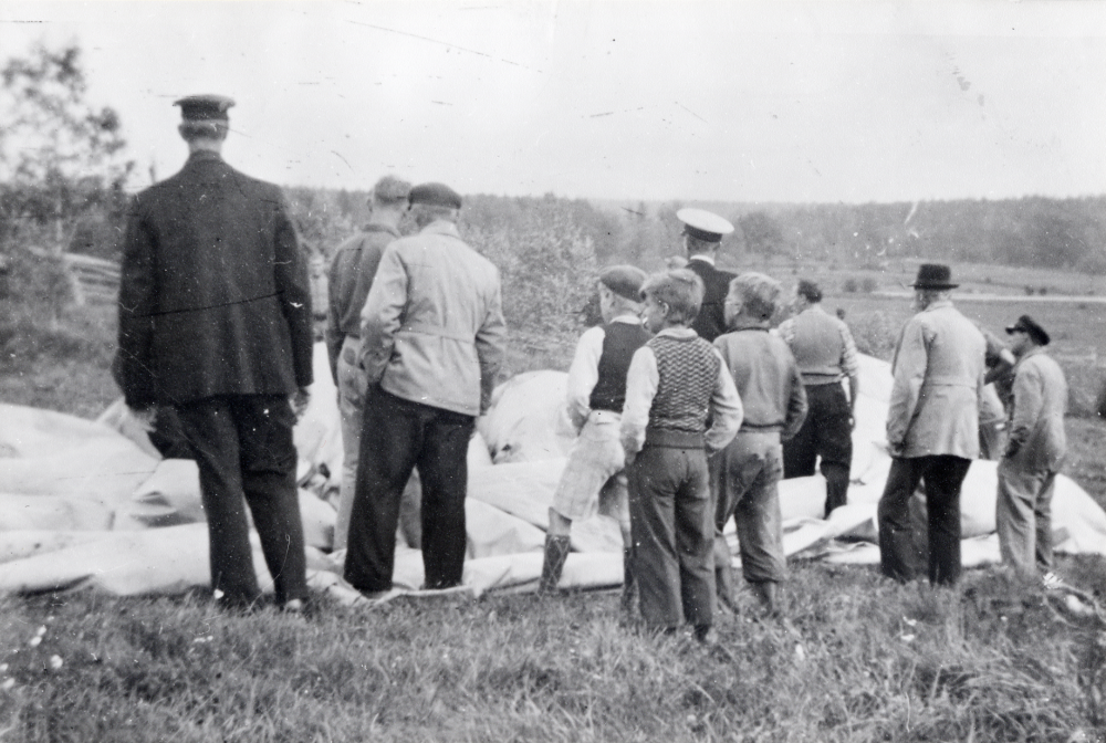 En spärrballong från England har landat vid Hall i Kårsta. En mängd män och småpojkar har samlats kring ballongen. En polis finns på plats samt en mörkklädd man med uniformsmössa?
