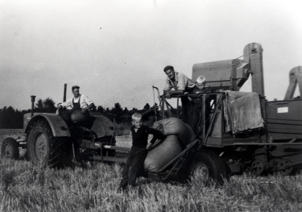 Skördetröska från Väsby gård vid Solgården i Vallentuna.  Sven-Erik Karlsson, Väsby gårds arrendator, sitter vid ratten på traktorn, en Diesel-Ross.  På skördetröskan, en 5 fot Munktell-tröska, står Sven Liljeholtz, som ofta hjälpte till vid gården.  Sven-Erik arrenderade även mark här vid Solgården, där man denna sensommar skördade vete.  Pojken på tröskan är Maj och Sven-Eriks son Mats, pojken på marken heter ev Kjell