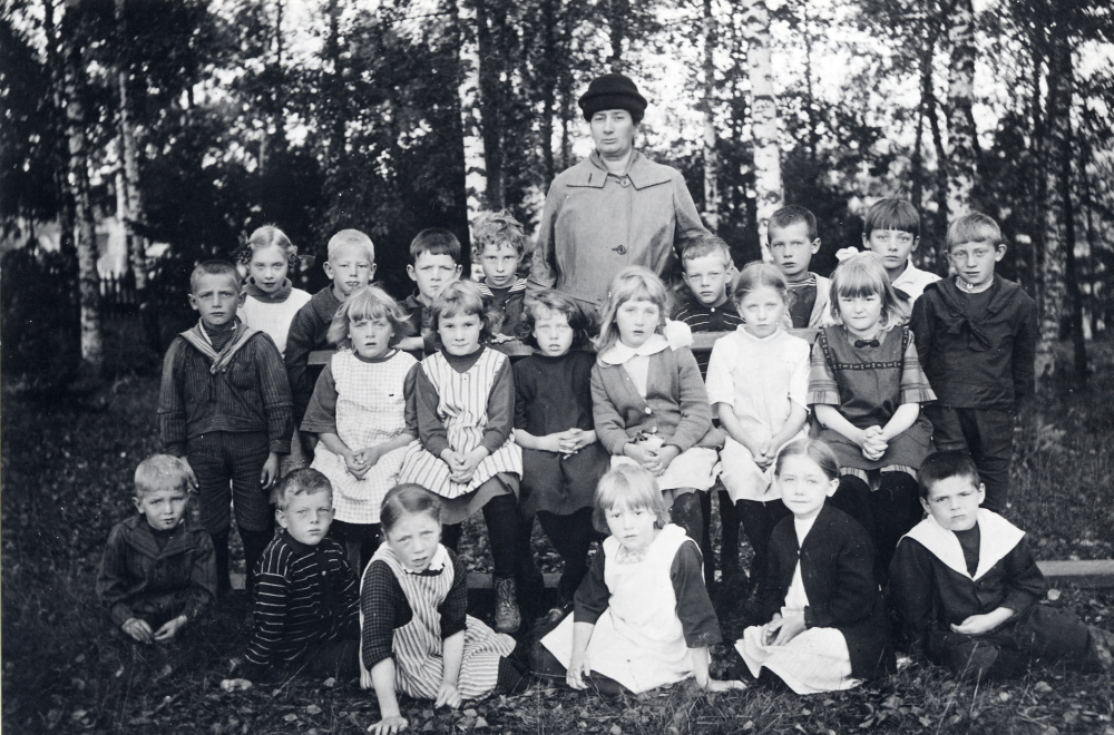 Lärarinnan Anna Gauffin omgiven av skolbarnen i Hjälmsta skola vid Gamla Missionkyrkan vid Olsborgsvägen. Bakre raden fr v: Nils ?, nr 3 Arne Lundqvist, nr 4 Axel Nordqvist, nr 6 Ivan Bergman, nr 7 Rickard Jansson, Andra raden fr v nr 1 Bertil Blom, nr 2 Karin Larsson, nr 3 Marianne Härje, (Orup), nr 4 Anna-Britta Fryckstedt, (Ekström) nr 6 Nancy Olofsson. Tredje raden fr v nr 5 Greta? Gustavsson, och Anders ?